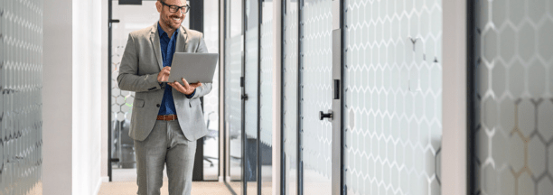 Professional man wearing glasses holding a laptop smiles and walks down hallway at work.
