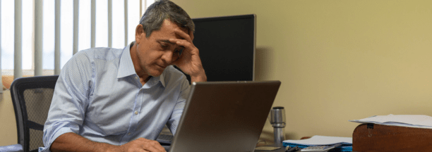 Minority man at work looking depressed while sitting at his desk on a laptop.