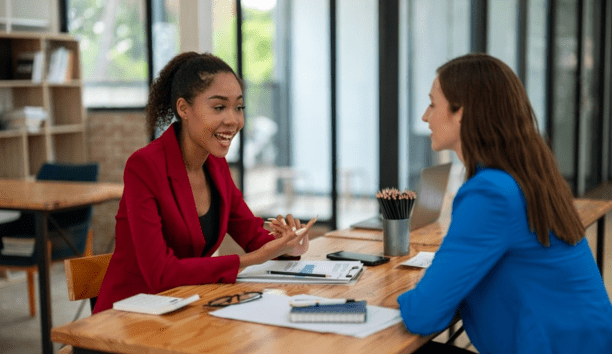 Two female employees discuss work facing each other at a table. 