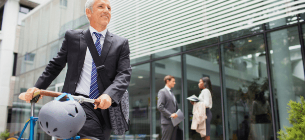 Man bikes to work with other professionals behind him in front of office building.