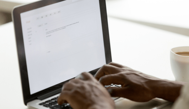 Hands of a person on a laptop keyboard writing an email with a cup of coffee next to them. 