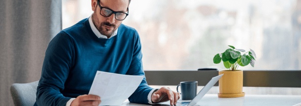 Man in blue sweater with glasses looks down at paper in his hand while sitting at a desk working.