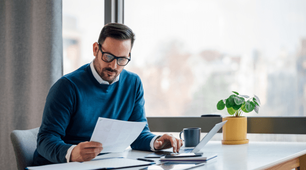 Man in blue sweater with glasses looks down at paper in his hand while sitting at a desk working.