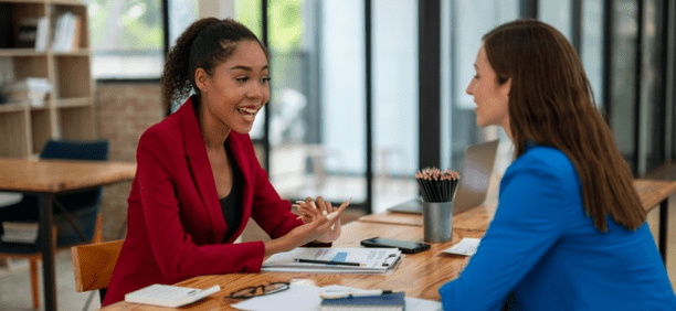 Two female employees discuss work facing each other at a table.