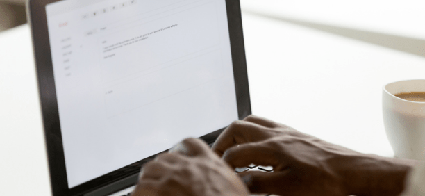 Hands of a person on a laptop keyboard writing an email with a cup of coffee next to them.
